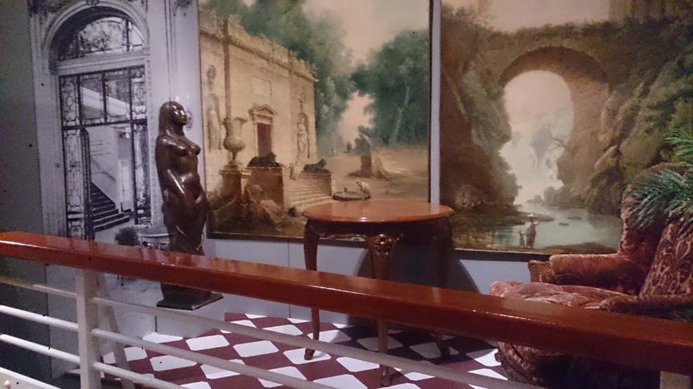 A statue, table and armchair in front of wallpaintings showing scenes of historical ruins.