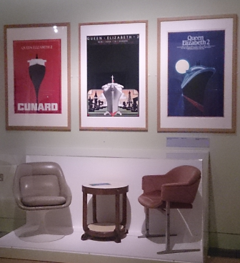 Two chairs and a side table on display beneath promotional posters of the QE2